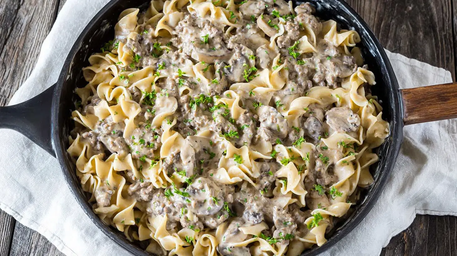 Hearty ground beef stroganoff in a dark skillet with creamy pasta and mushrooms on a rustic wooden background.
