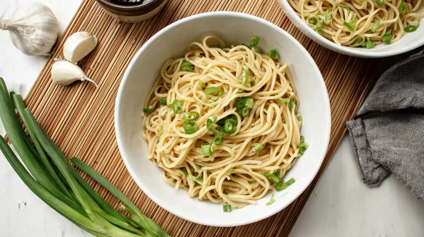 Overhead view of two bowls of savory garlic noodles garnished with fresh green onions, surrounded by whole garlic cloves and uncooked scallions on a rustic bamboo mat.